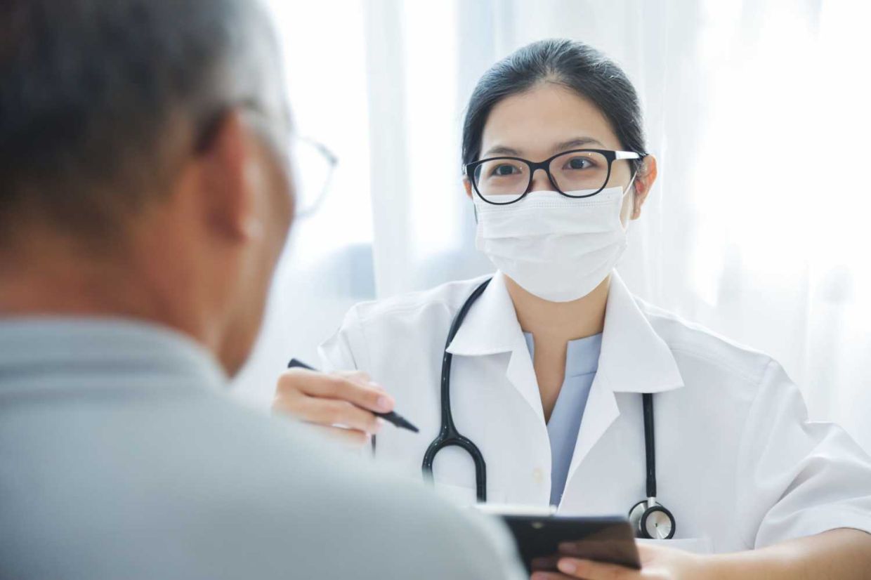 Young Asian Female Doctor with protective mask talking to senior man patient in medical office.